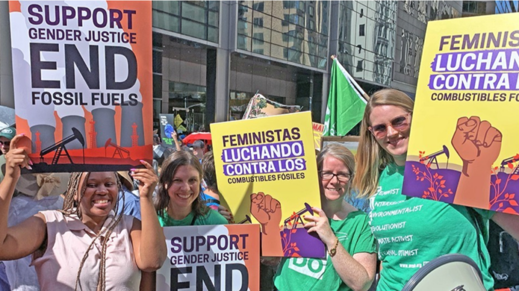 Image description: On a sunny day in New York, four feminists with banners pose at the march to demand an end to fossil fuels. 